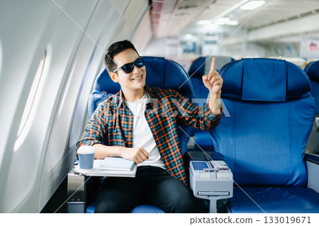 Happy young traveler celebrating victory with a fist pump on the airplane. Business success, achievement, and excited travel journey. Win, style, phone, tech. Happy young traveler celebrating victory with a fist pump on the airplane. Business success, achievement, and excited travel journey. Win, style, phone, tech. 133019671