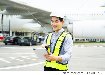 Ground crew engineer in safety vest and hard hat using walkie talkie while checking documents, perfect for aviation, maintenance, 133019712