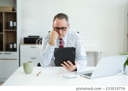 Senior businessman in formal attire using tablet and drinking coffee at modern office desk, working on finance, planning, or digital consulting tasks. Senior businessman in formal attire using tablet and drinking coffee at modern office desk, working on finance, planning, or digital consulting tasks. 133019759