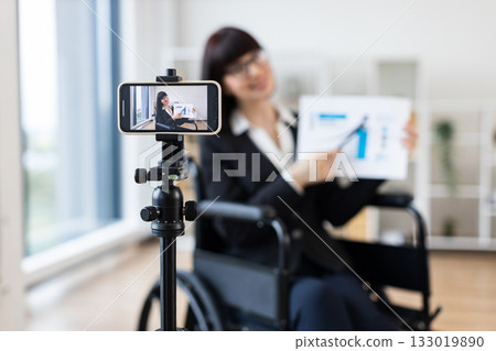 Caucasian woman in wheelchair blogs on camera about business using charts and graphs. Indoor setting with focus on professional blogging and business concepts. Caucasian woman in wheelchair blogs on camera about business using charts and graphs. Indoor setting with focus on professional blogging and business concepts. 133019890
