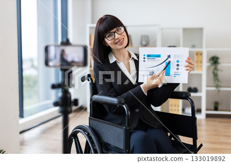 Caucasian woman in wheelchair holding chart showing business data while blogging on camera 133019892