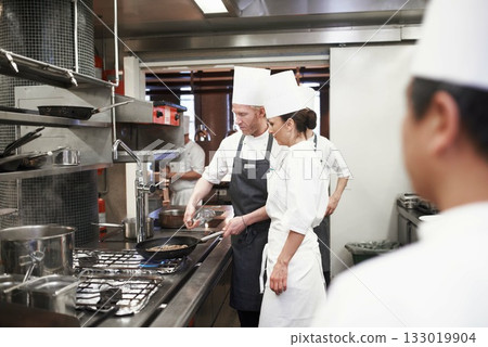 Chef, teamwork and people with frying pan in kitchen, catering service and prepare food for fine dining. Professional, cooking and man training woman, saute and cook add oil to meat in restaurant. 133019904