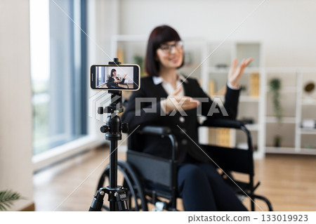 Caucasian woman in business attire sits in wheelchair recording informative business blog on smartphone. Modern interior, professional appearance, creating content for business communication. 133019923