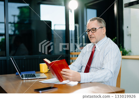 Mature Businessman using laptop while sitting at home. man sitting in kitchen and doing market research working Mature Businessman using laptop while sitting at home. man sitting in kitchen and doing market research working 133019926