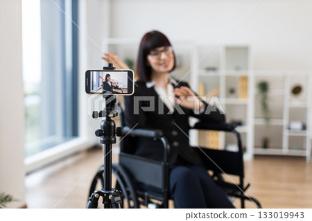 Caucasian woman in business attire sits in wheelchair recording informative business blog on smartphone. Modern interior, professional appearance, creating content for business communication. 133019943