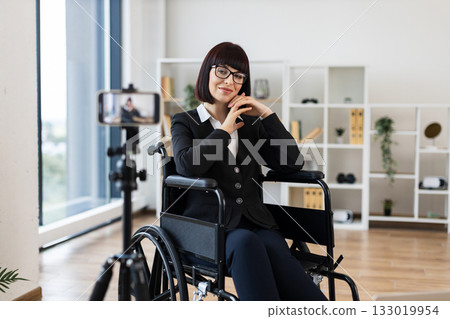 Caucasian woman wearing formal suit, sitting in wheelchair, recording business blog on smartphone camera in modern bright office. Represents entrepreneurship, determination, accessibility. 133019954