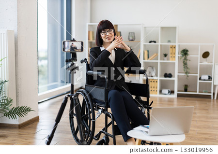 Caucasian woman wearing formal black attire sits in wheelchair in modern office, recording business-related video content using tripod-mounted camera and laptop, audience through relevant explanations 133019956