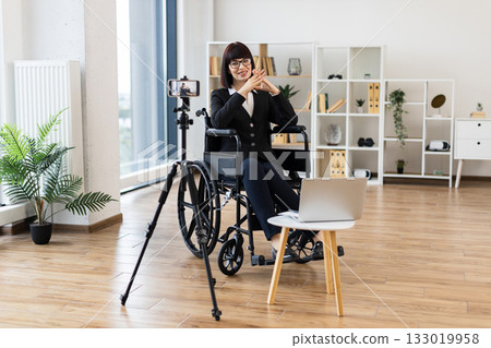Caucasian woman wearing formal black attire sits in wheelchair in modern office, recording business-related video content using tripod-mounted camera and laptop, audience through relevant explanations Caucasian woman wearing formal black attire sits in wheelchair in modern office, recording business-related video content using tripod-mounted camera and laptop, audience through relevant explanations 133019958