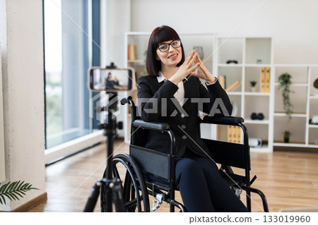 Caucasian woman wearing formal clothes sits in wheelchair recording business blog onto camera at office. Motivational and entrepreneurial vibes emphasizing inclusion, diversity, professional insights. 133019960
