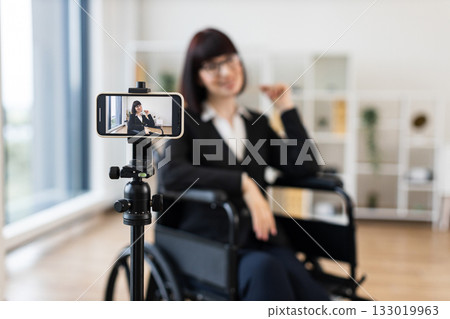 Caucasian woman wearing formal clothes sits in wheelchair recording business blog onto camera at office. Motivational and entrepreneurial vibes emphasizing inclusion, diversity, professional insights. 133019963