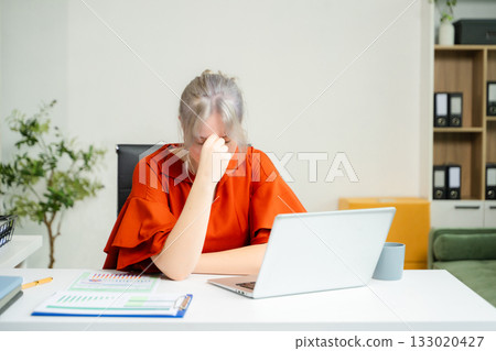 Overwhelmed young woman stressed at her desk with a laptop and reports in a modern office. Concept of burnout, workplace pressure 133020427