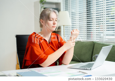 Young woman touching her neck in pain while working on laptop in bright office. Concept of workplace discomfort, poor posture, stress, and remote job fatigue. 133020430