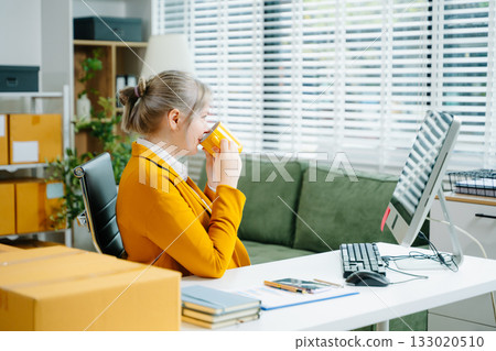 Young SME businesswoman in yellow blazer using smartphone and computer for online shopping and work in modern office. Digital tech, freelance, real people. 133020510