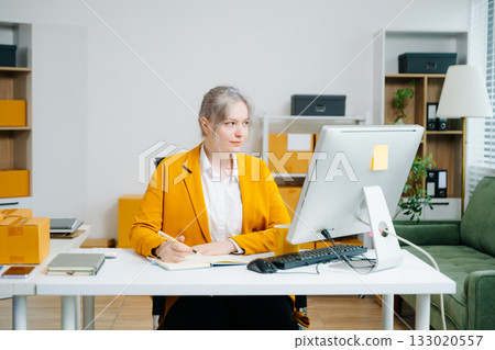Young SME businesswoman in yellow blazer using smartphone and computer for online shopping and work in modern office. Digital tech, freelance, real people. Young SME businesswoman in yellow blazer using smartphone and computer for online shopping and work in modern office. Digital tech, freelance, real people. 133020557