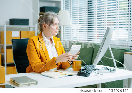 Young SME businesswoman in yellow blazer using smartphone and computer for online shopping and work in modern office. Digital tech, freelance, real people. 133020563
