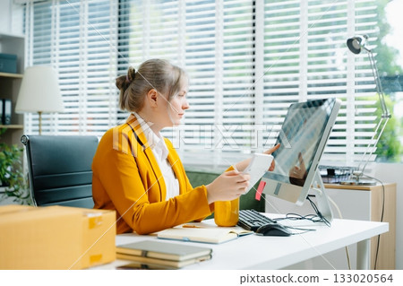 Young SME businesswoman in yellow blazer using smartphone and computer for online shopping and work in modern office. Digital tech, freelance, real people. 133020564