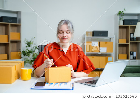 Young Caucasian woman entrepreneur packing boxes for shipping in home office, working with laptop, clipboard, and parcels. E-commerce and small business concept. Young Caucasian woman entrepreneur packing boxes for shipping in home office, working with laptop, clipboard, and parcels. E-commerce and small business concept. 133020569