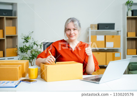 Young Caucasian woman celebrating a successful online business at home, surrounded by boxes and laptop. E-commerce, entrepreneur, and shipping concept 133020576