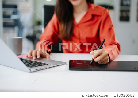 Happy Asian woman creating digital content on laptop and drawing tablet in modern office. Happy Asian woman creating digital content on laptop and drawing tablet in modern office. 133020592