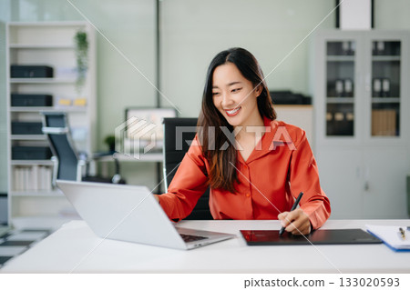 Happy Asian woman creating digital content on laptop and drawing tablet in modern office. 133020593