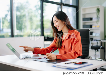 Happy Asian woman creating digital content on laptop and drawing tablet in modern office. Happy Asian woman creating digital content on laptop and drawing tablet in modern office. 133020594
