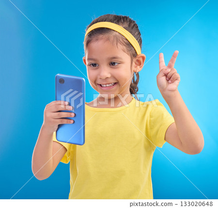 Phone, girl kid and peace sign selfie in studio isolated on a blue background. Technology, v hand emoji and smile of child with mobile smartphone to take pictures for happy memory or social media. Phone, girl kid and peace sign selfie in studio isolated on a blue background. Technology, v hand emoji and smile of child with mobile smartphone to take pictures for happy memory or social media. 133020648