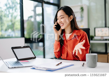 Happy Asian businesswoman speaking on phone at desk in a bright office. Concept of client communication Happy Asian businesswoman speaking on phone at desk in a bright office. Concept of client communication 133021087