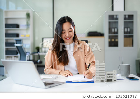 Confident Asian businesswoman smiling while presenting a modern building model in office 133021088