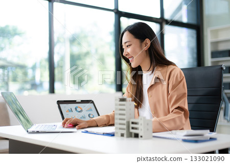 Asian woman analyzing sales data on tablet with real estate model on desk. Ideal for finance, property investment, marketing, and smart business strategy concepts. Asian woman analyzing sales data on tablet with real estate model on desk. Ideal for finance, property investment, marketing, and smart business strategy concepts. 133021090