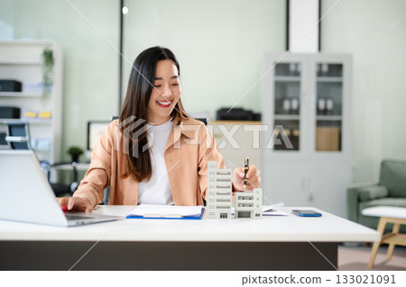 Young real estate agent worker working with laptop and tablet at table in office and small house beside it. Young real estate agent worker working with laptop and tablet at table in office and small house beside it. 133021091
