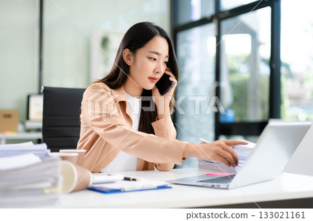 Woman holding coffee and talking on the phone while working at a cluttered office desk with laptop, files, and paperwork 133021161