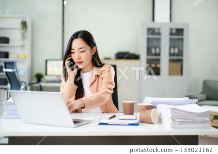 Woman holding coffee and talking on the phone while working at a cluttered office desk with laptop, files, and paperwork 133021162