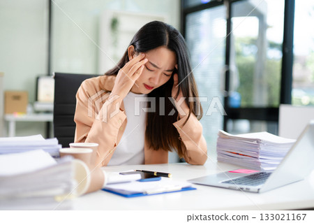 Asian businesswoman suffering headache from stress and work pressure. Holding head in pain at office desk Asian businesswoman suffering headache from stress and work pressure. Holding head in pain at office desk 133021167