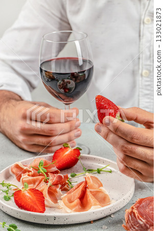 Man holding glass of red wine and strawberry with prosciutto appetizer on plate, close up Man holding glass of red wine and strawberry with prosciutto appetizer on plate, close up 133021873