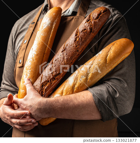 Baker holding assortment of fresh baguettes on dark background, copy space Baker holding assortment of fresh baguettes on dark background, copy space 133021877