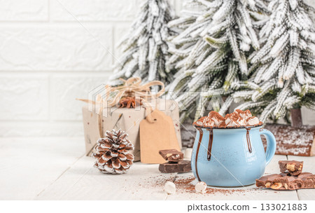 Cup of hot chocolate with marshmallows in blue mug on wooden table with Christmas decorations, pine cone, gift box and snowy fir trees, cozy winter holiday scene with copy space Cup of hot chocolate with marshmallows in blue mug on wooden table with Christmas decorations, pine cone, gift box and snowy fir trees, cozy winter holiday scene with copy space 133021883