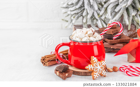 Red mug filled with hot chocolate and marshmallows, candy cane, gingerbread cookie and chocolate pieces on light background with snowy Christmas tree, festive composition with copy space 133021884