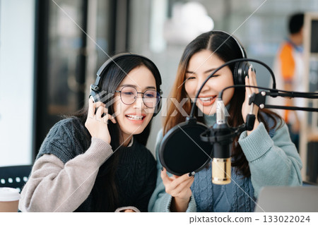 Young woman and Asian woman wearing headphones and doing a live podcast for their channel, communication for radio podcast and technology concept Young woman and Asian woman wearing headphones and doing a live podcast for their channel, communication for radio podcast and technology concept 133022024