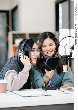 Young woman and Asian woman wearing headphones and doing a live podcast for their channel, communication for radio podcast and technology concept Young woman and Asian woman wearing headphones and doing a live podcast for their channel, communication for radio podcast and technology concept 133022027