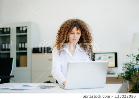 woman is sitting at a desk with a laptop and a cell phone while holding coffee at office woman is sitting at a desk with a laptop and a cell phone while holding coffee at office 133022080