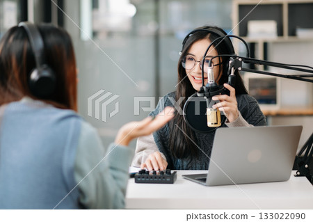 Young woman and Asian woman wearing headphones and doing a live podcast for their channel, communication for radio podcast and technology concept Young woman and Asian woman wearing headphones and doing a live podcast for their channel, communication for radio podcast and technology concept 133022090