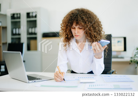 woman is sitting at a desk with a laptop and a cell phone while holding coffee at office woman is sitting at a desk with a laptop and a cell phone while holding coffee at office 133022132