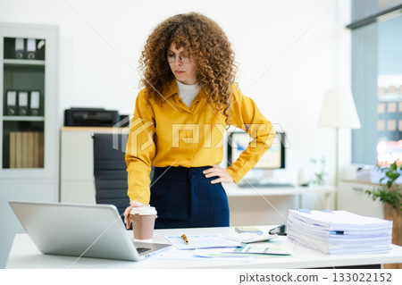 Curly haired businesswoman writing on paperwork at desk with laptop, documents, and coffee. Concept 133022152