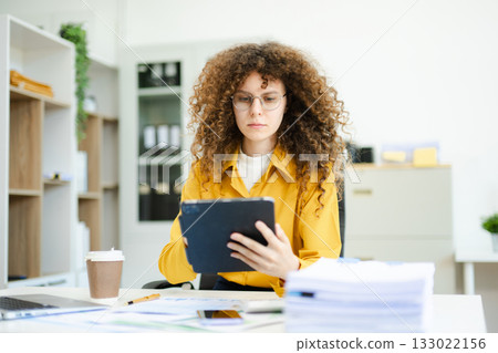 Confident businesswoman analyzing data on tablet at modern office desk with coffee, documents, and laptop. Concept of digital work Confident businesswoman analyzing data on tablet at modern office desk with coffee, documents, and laptop. Concept of digital work 133022156