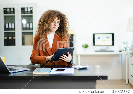 Focused businesswoman working on laptop while holding tablet at desk with documents and coffee. Concept of productivity, multitasking Focused businesswoman working on laptop while holding tablet at desk with documents and coffee. Concept of productivity, multitasking 133022227