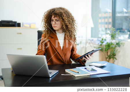 Focused businesswoman working on laptop while holding tablet at desk with documents and coffee. Concept of productivity, multitasking 133022228