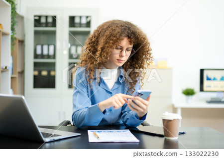 woman is sitting at a desk with a laptop and a cell phone while holding coffee at office woman is sitting at a desk with a laptop and a cell phone while holding coffee at office 133022230