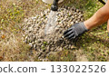 Hand of a worker using a hose to spray water on gravel pile in a garden, showcasing landscaping techniques and maintenance practices 133022526