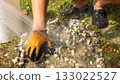 Hand in protective glove is washing gravel surface with water from a bucket, showcasing outdoor cleaning process and maintenance efforts 133022527