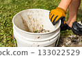 Person wearing gloves is lifting a dirty bucket filled with gravel and debris, showcasing outdoor cleanup efforts in a natural environment 133022529